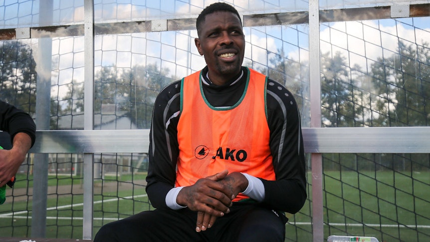 Ex-Gladbacher Mo Idrissou sitz am 10. Oktober 2020 im orangenen Leibchen beim Spiel gegen den Hunfelder SV auf der Ersatzbank beim SC Viktoria Griesheim und lächelt.