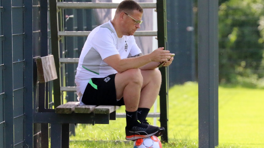 Gladbach-Manager Max Eberl sitzt auf einer Bank, derweil schaut er auf sein Smartphone. Seine Füße ruhen auf einem Fußball.