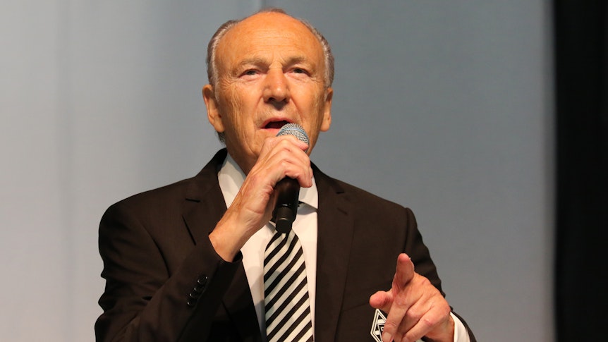 Borussias Präsident Rolf Königs bei der Mitgliederversammlung am 10. August 2021 im Gladbacher Stadion. Der 79-Jährige hält in der rechten Hand ein Mikro, mit der linken Hand deutet er in Richtung Zuhörer.