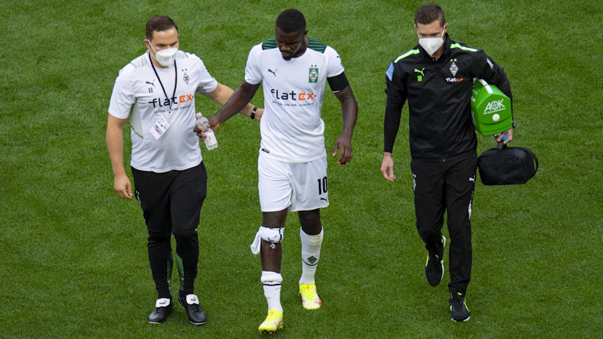 Gladbach-Stürmer Marcus Thuram (Mitte) humpelt beim verlorenen Rheinland-Duell (0:4) der Borussia in Leverkusen (21. August 2021) frühzeitig verletzt vom Platz. Er wird von zwei Betreuern, die eine Maske tragen, begleitet.