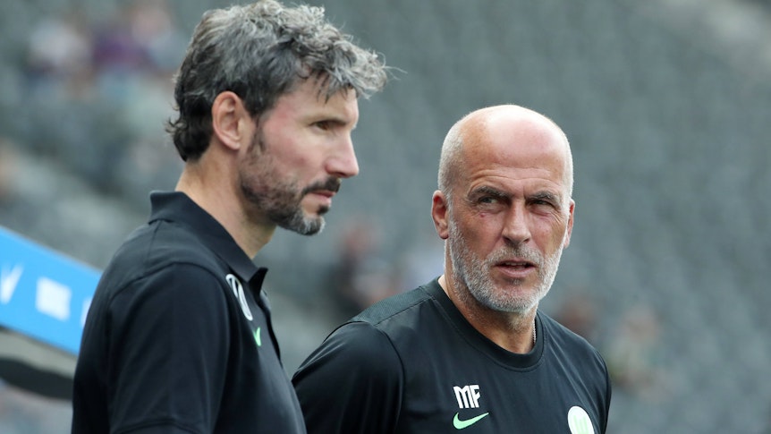 Ex-Gladbach-Trainer Michael Frontzeck (r.) arbeitet aktuell als Co-Trainer unter Mark van Bommel (l.) beim Fußball-Bundesligisten VfL Wolfsburg. Auf diesem Foto sind die beiden am 21. August 2021 im Berliner Olympiastadion zu sehen. Frontzeck blickt zu van Bommel rüber.