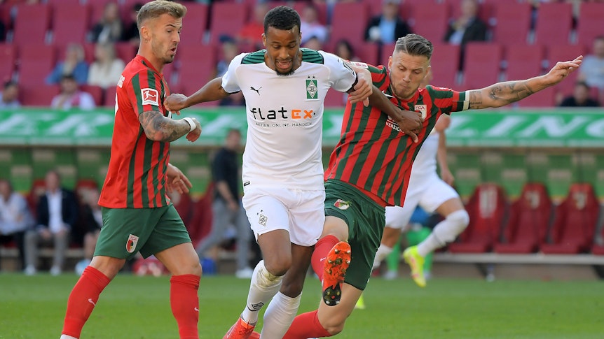 Alassane Plea von Borussia Mönchengladbach versucht seine beiden Augsburger Gegenspieler mit ausgestreckten Armen von sich wegzuhalten im Spiel gegen den FCA am 18. September 2021.