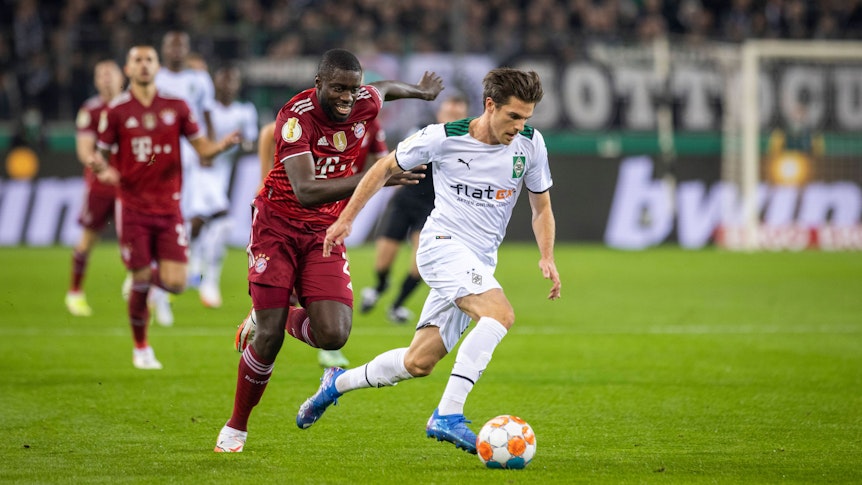 Verteidiger Dayot Upamecano (l.) von Bayern München läuft Offensivspieler Jonas Hofmann (r.) von Borussia Mönchengladbach im DFB-Pokalspiel am 27. Oktober 2021 im Borussia-Park hinterher. Hofmann sprintet mit dem Ball am rechten Fuß dem Franzosen davon.