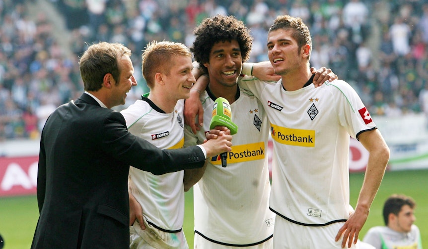 Stadionssprecher Torsten Knippertz im Gespräch mit Marco Reus, Dante und Roman Neustädter (v.l.n.r.) im Borussia-Park. Das Foto stammt vom 28. April 2012, die drei Spieler haben Borussia Mönchengladbach am Ende der Saison 2011/2012 verlassen. Reus, Dante und Neustädter umarmen sich, derweil hält Knippertz ihnen ein Mikrofon hin.