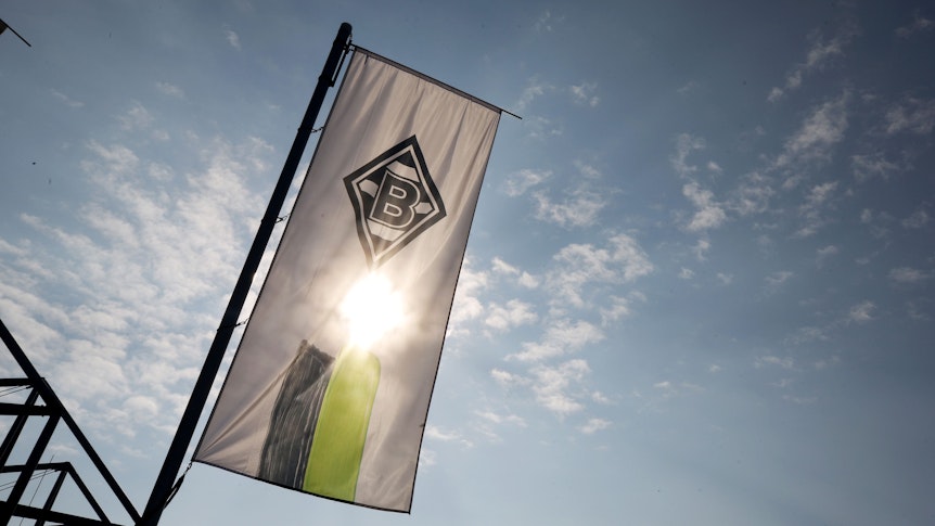 Eine Fahne mit dem Logo von Borussia Mönchengladbach weht am Borussia-Park. Auf dem Symbolfoto scheint die Sonne, sie wird von der Fahne allerdings verdeckt.