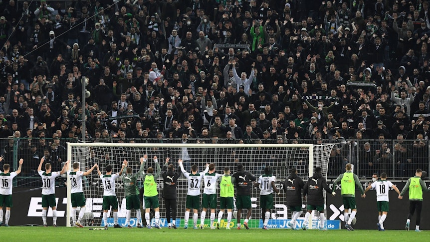 Die Spieler von Borussia Mönchengladbach feiern am Samstag (12. März 2022) gemeinsam mit den Fans den Heimsieg im Abstiegskampf gegen Hertha BSC. Die Gladbach-Spieler bilden eine Kette und heben gemeinsam die Arme.
