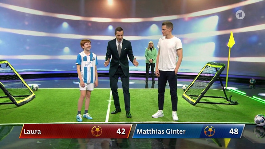 Moderator Kai Pflaume (Mitte) mit Nationalspieler Matthias Ginter (r.) von Borussia Mönchengladbach und der 13-jährigen Laura (l.) in der ARD-Sendung „Klein gegen Groß – Das unglaubliche Duell“ am 12. März 2022. Pflaume macht eine Geste, Ginter und Laura lachen.