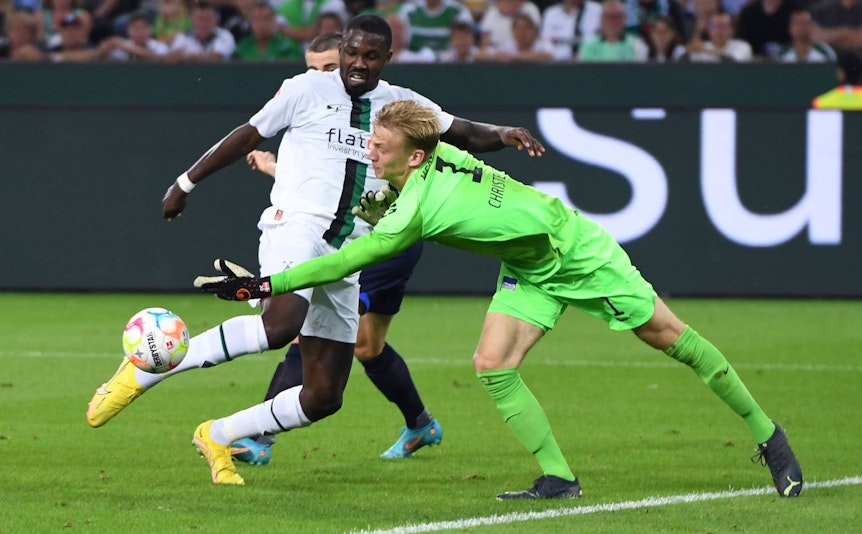 Gladbach-Stürmer Marcus Thuram (l.) im Duell mit Hertha-Schlussmann Oliver Christensen (r.) während der Bundesliga-Partie am Freitag (19. August 2022) im Borussia-Park. Beide schauen auf den Ball.