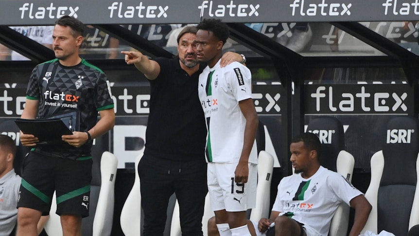 Trainer Daniel Farke von Borussia Mönchengladbach gibt Nathan Ngoumou Anweisung vor dessen Einwechslung am 4. September 2022 gegen Mainz 05. Farke hat den Arm um Ngoumous Schulter gelegt und zeigt mit dem anderen Arm auf das Spielfeld. Links steht Co-Trainer Edmund Riemer, im Hintergrund sitzt Alassane Plea auf der Bank.