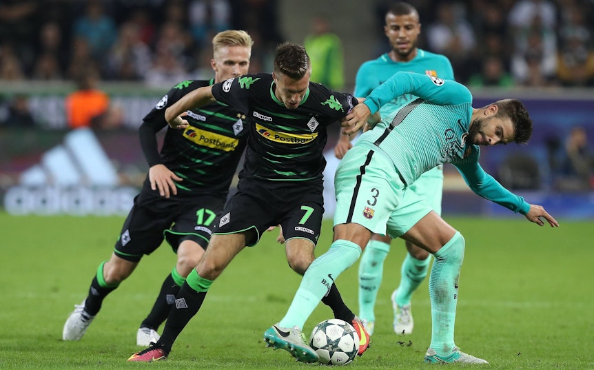 Gladbach-Profi Patrick Herrmann (Mitte) im Champions-League-Duell am 28. September 2016 mit dem FC Barcelona im Borussia-Park. In dieser Szene befindet sich der Flügelflitzer im Zweikampf mit Abwehrspieler Gerard Piqué. Beide Spieler haben jeweils den Ball im Blick.
