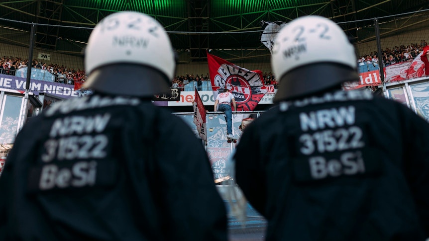 Polizeikräfte beobachten den Gästeblock im Borussia-Park. Dieses Symbol-Foto stammt vom Bundesliga-Derby zwischen Borussia Mönchengladbach und dem 1. FC Köln am 16. April 2022. Zu sehen sind zwei Polizisten in Einsatzmontur, sie tragen Schutzhelme und blicken hoch zu den FC-Fans.
