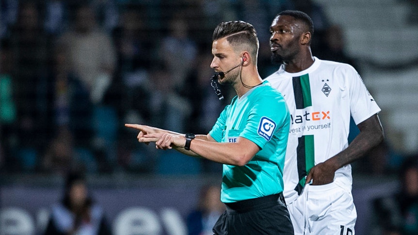 Schiedsrichter Daniel Schlager zeigt an, dass es eine VAR-Entscheidung im Spiel VfL Bochum gegen Borussia Mönchengladbach am 8. November 2022 gibt. Borussia-Spieler Marcus Thuram steht dabei hinter ihm.