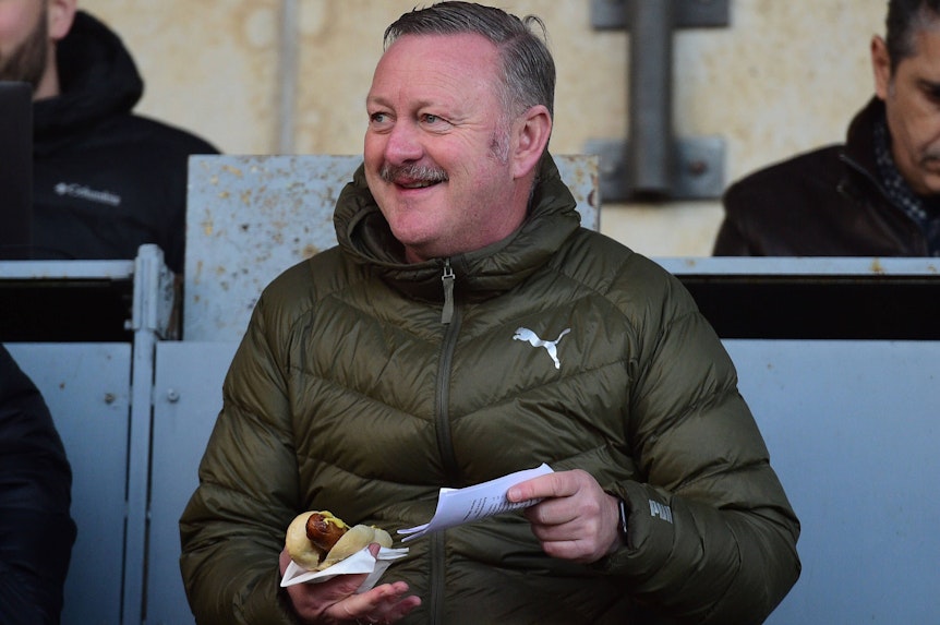 Da schmeckt dem Gladbach-Manager die Borussia-Wurst! Sportdirektor Roland Virkus hat sich am Freitag (18. November 2022) die Partie der U23 in der Regionalliga West gegen Schalke 04 im Rheydter Grenzlandstadion angeschaut. Virkus hält eine Bratwurst im Bröchten in seinen rechten Hand, während er auf der Tribüne sitzt.