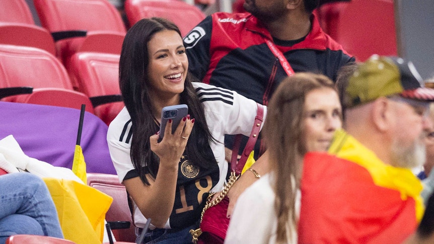 Sky-Moderatorin Laura Winter, hier zu sehen am 1. Dezember 2022 während des WM-Spiels der deutschen Nationalmannschaft gegen Costa Rica in Doha, ist mit DFB-Spieler und Gladbach-Star Jonas Hofmann verheiratet. Winter trägt ein Deutschland-Trikot und hält ein Smartphone in ihrer rechte Hand. Sie lächelt zudem.