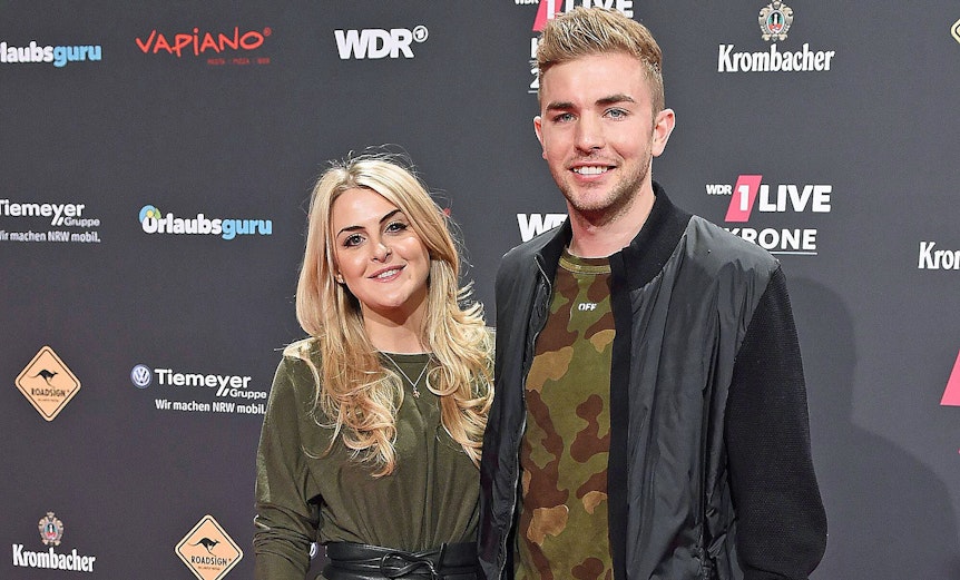ZDF-Experte und Gladbach-Profi Christoph Kramer (r.) mit seiner Frau Celina (l.) bei der „1LIVE Krone“ am 7. Dezember 2017 in der Jahrhunderthalle in Bochum. Die beiden lächeln, hinter ihnen ist eine Werbewand zu sehen.