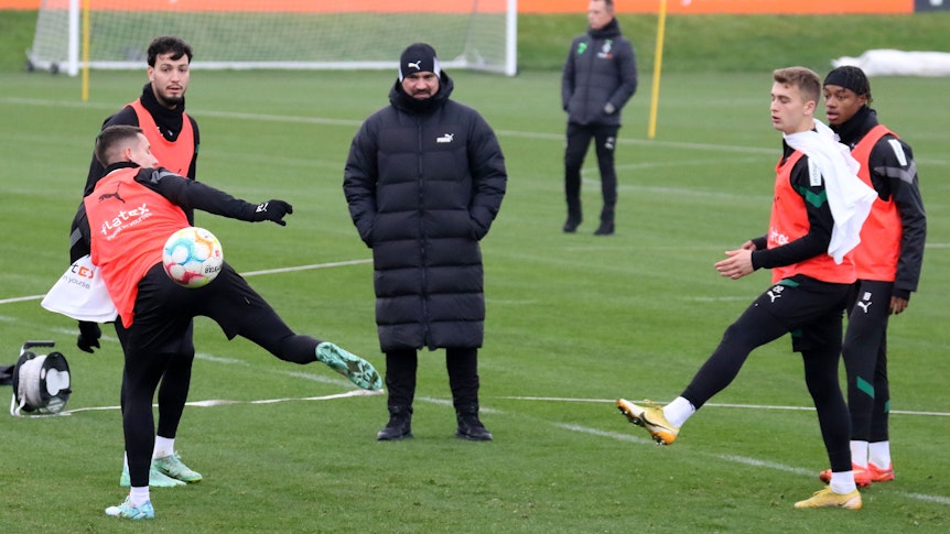 Trainings von Borussia Mönchengladbach im Borussia-Park am 22. Dezember 2022. Trainer Daniel Farke (M.) schaut seinen Spielern Conor Noß, Ramy Bensebaini, Oscar Fraulo und Yvandro Borges Sanches (v. l. n. r.) zu. Farke trägt eine lange Winterjacke und eine Mütze.