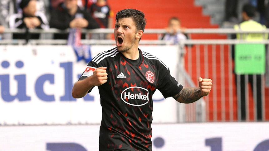Dawid Kownacki von Fortuna Düsseldorf brüllt nach einem Tor den Fans auf der Tribüne zu und jubelt mit nach vorne gestreckten Armen. Der polnische Stürmer trägt das rot-schwarze Auswärtstrikot von Fortuna.