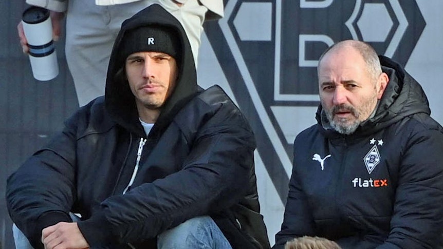 Gladbach-Torwart Yann Sommer (l.) sitzt am Samstag (7. Januar 2023) neben Co-Trainer Oliver Neuville (r.) und beobachtet das Testspiel seiner Team-Kollegen gegen Drittligist Oldenburg. Sommer hat eine Mütze auf.