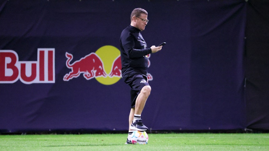 Ex-Gladbach-Manager Max Eberl ist mit seinem neuen Klub RB Leipzig im Trainingslager in Abu Dhabi. Auf diesem Foto ist der 49-Jährige am 3. Januar 2023 zu sehen. Eberl hält in der rechten Hand sein Smartphone, auf das er blickt. Sein rechter Fuß steht auf einem Ball.