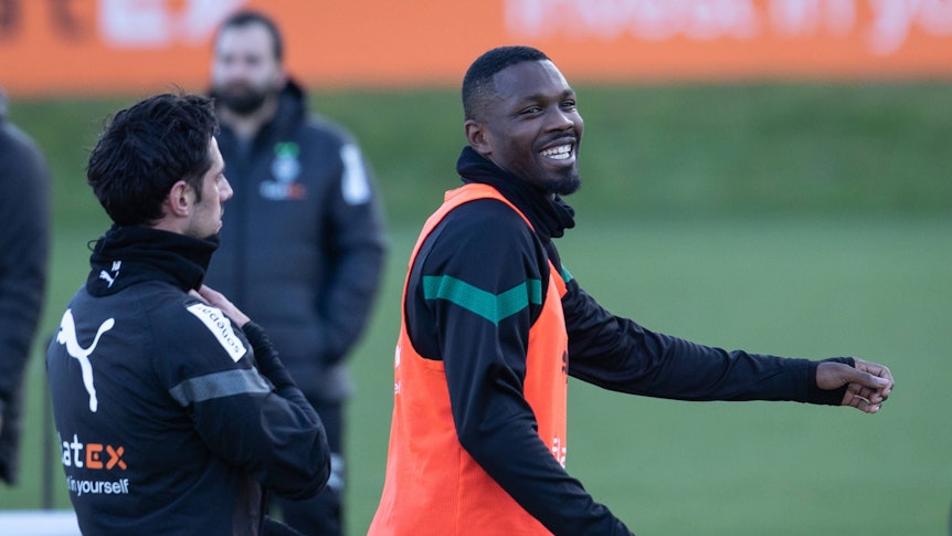 Marcus Thuram steht in Trainingskleidung und einem roten Leibchen am 6. Januar 2023 auf dem Trainingsplatz von Borussia Mönchengladbach. Der Fußballer lacht nach einem Gespräch mit Borussia-Teamkollege Lars Stindl.