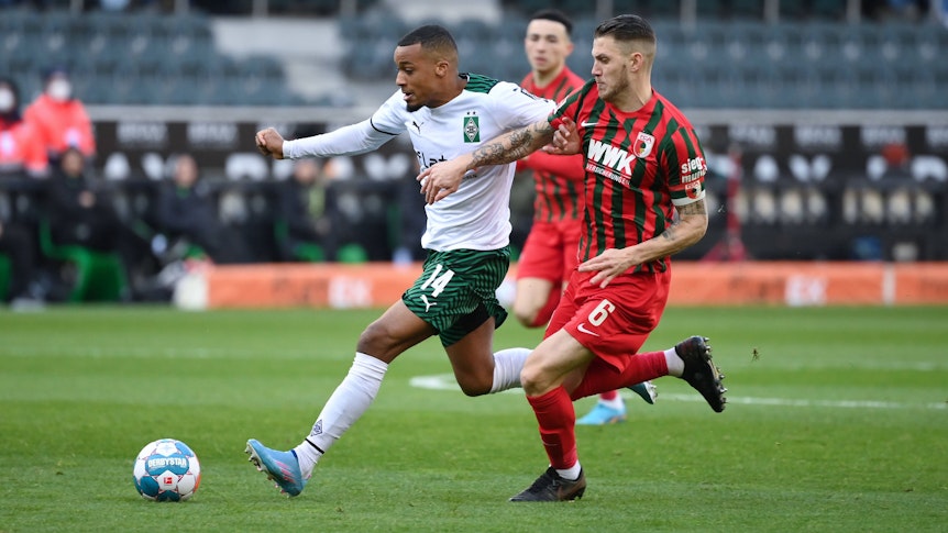 Alassane Plea (l.) von Borussia Mönchengladbach bestreitet einen Zweikampf mit Jeffrey Gouweleeuw vom FC Augsburg. Plea läuft dem Ball entgegen und trägt ein weiß-grünes Borussia-Trikot. Gouweleeuw verfolgt seinen Gegenspieler in einem rot-grünen Trikot.