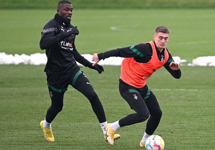 Gladbach-Stürmer Marcus Thuram (l.) im Duell mit Team-Kollege Oscar Fraulo (r.), hier zu sehen am Montag (23. Januar 2023) während einer Fohlen-Trainingseinheit im Borussia-Park. Fraulo trägt ein orangefarbenes Leibchen und hat den Ball vor sich.