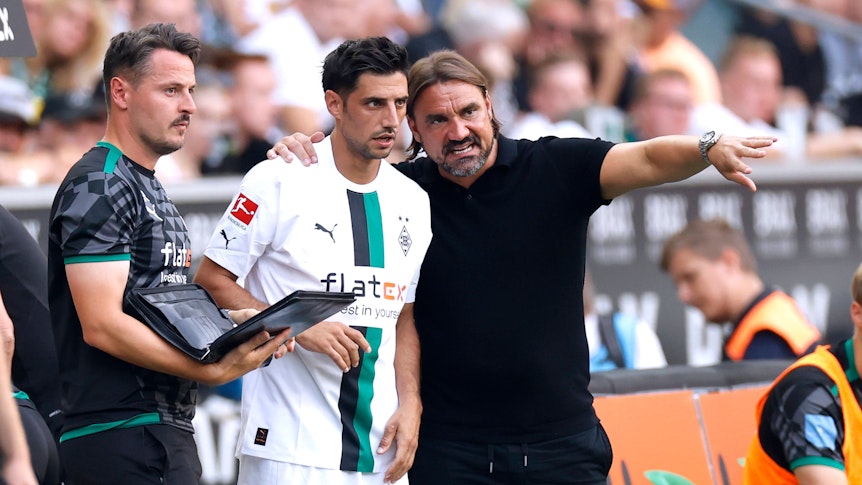 Daniel Farke (r.) legt seinen Arm um Lars Stindl (M.) und gibt dem Kapitän von Borussia Mönchengladbach vor einer Einwechslung taktische Anweisungen. Farke macht zudem eine Geste mit seinem linken Arm. Neben Farke und Stindl steht Co-Trainer Edmund Riemer und hält eine Mappe in der Hand.