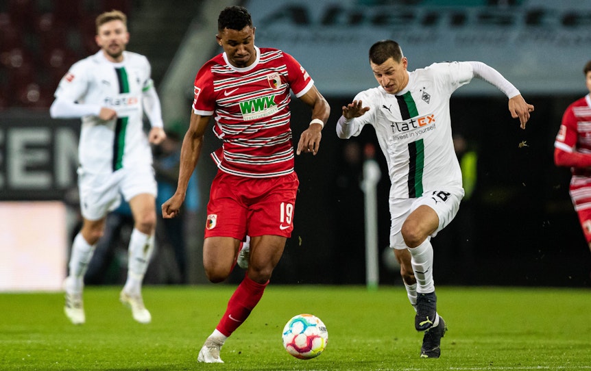 Gladbach-Verteidiger Stefan Lainer (r.) im Duell mit Felix Uduokhai (l.) vom FC Augsburg während des Bundesliga-Duells am Mittwoch (25. Januar 2023) in der WWK-Arena. Beide Spieler haben den Ball im Blick und sprinten.