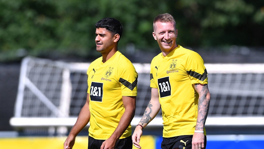 Mo Dahoud (l.) steht im Trainingslager von Borussia Dortmund neben seinem Teamkollegen Marco Reus. Beide tragen auf dem Rasen Trainingskleidung des BVB und lachen.