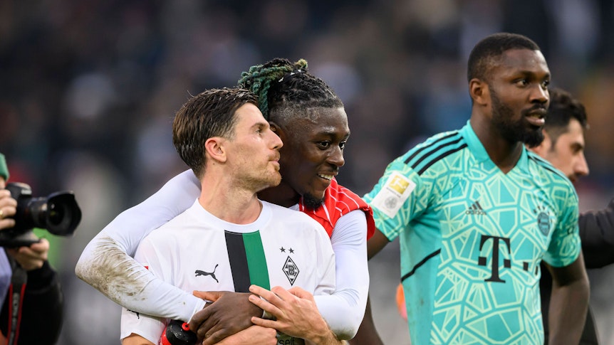 Manu Koné steht nach dem Bundesliga-Sieg von Borussia Mönchengladbach gegen den FC Bayern München am 18. Februar 2023 hinter seinem Teamkollegen Jonas Hofmann und legt seine Arme freundschaftlich um ihn. Marcus Thuram (r.) steht im Bayern-Trikot von Yann Sommer neben dem Gladbach-Duo auf dem Rasen im Borussia-Park.
