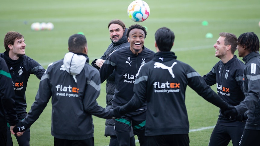 Ein bisschen Spaß muss sein! Gute Laune bei Borussia nach dem Heim-Coup gegen Bayern München. Die Gladbach-Profis, hier zu sehen am 16. Februar im Borussia-Park, beginnen nun mit der Vorbereitung auf das Mainz-Spiel. Zu sehen ist Ngoumou beim Kopfball, der Franzose lächelt, er hält seinen Team-Kollegen, die einen Kreis gebildet haben, an den Händen. Im Hintergrund schaut Trainer Daniel Farke zu.