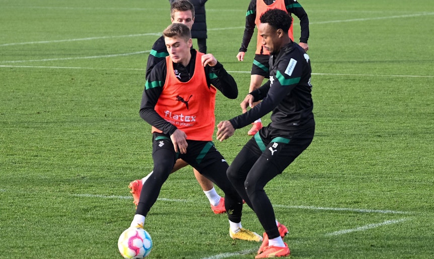 Kampf um den Ball zwischen den Gladbach-Talenten Oscar Fraulo (l.) und Nathan Ngoumou (r.) am 22. Dezember 2022 auf dem Trainingsgelände im Borussia-Park. Fraulo trägt ein Leibchen, beide Spieler sind auf den Ball fokussiert. Im Hintergrund schauen, Herrmann, Hofmann und Farke zu.