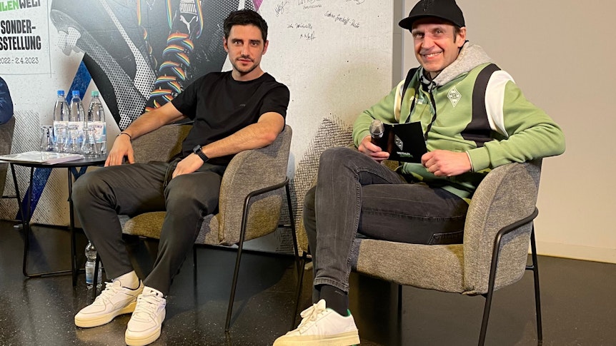 Gladbach-Kapitän Lars Stindl (l.) und Borussias Stadionsprecher Torsten Knippertz (r.) bei einer Diskussion (28. Februar 2023) zu den Themen Diskriminierung und Antidiskriminierung in der Fohlen-Welt im Borussia-Park. Beide schauen in die Kamera, Knippertz trägt eine Kappe.