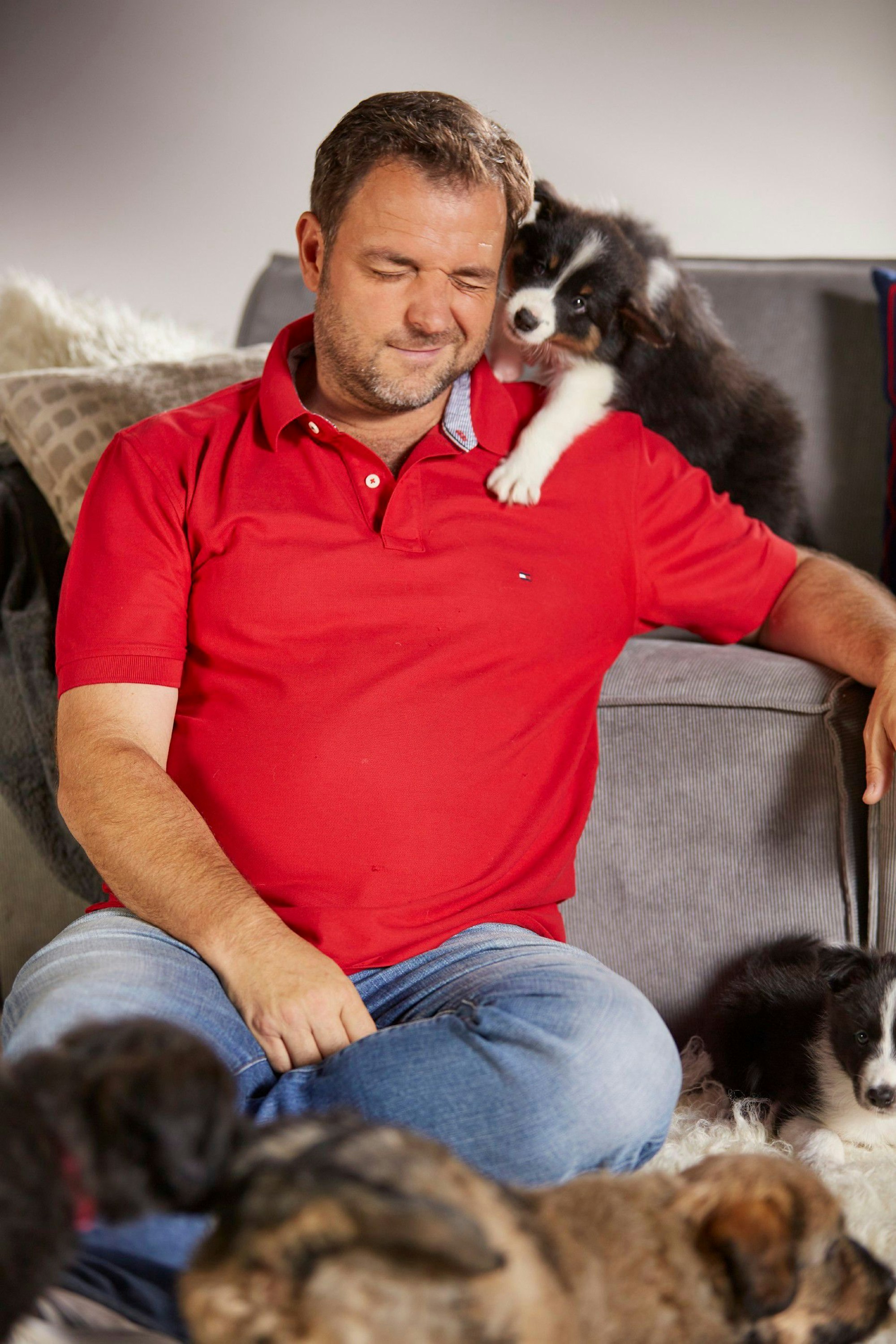 Hundeprofi Martin Rütter sitzt vor einer Couch und kuschelt mit einigen Hundewelpen. 