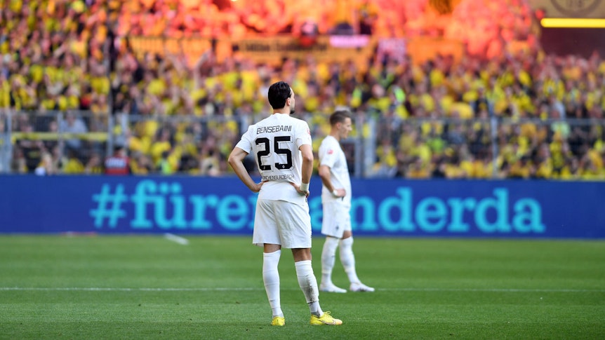 Ramy Bensebaini von Borussia Mönchengladbach ist nach einem Gegentreffer am 13. Mai 2023 bei Borussia Dortmund konsterniert. Er steht mit in die Hüften gestemmten Armen auf dem Rasen, im Hintergrund feiern die BVB-Fans auf der Südtribüne.
