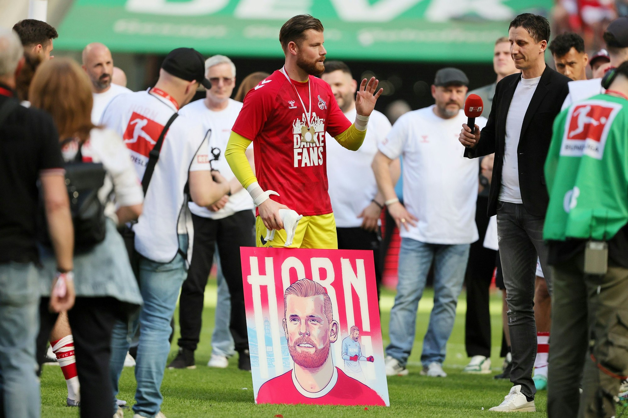 Tim Horn steht auf dem Platz, neben im Christian Keller. Im Hintergrund Steffen Baumgart. Vor Horn steht ein großes Bild, das ihn und seinen Nachnamen zeigt.