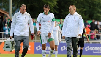 Florian Neuhaus (M.) verlässt gemeinsam mit Physiotherapeut Dirk Müller (l.) und Mannschaftsarzt Dr. Ralf Doyscher beim Testspiel von Borussia Mönchengladbach gegen den FC Ingolstadt am 26. Juli 2023 den Platz. Neuhaus' linker Oberschenkel ist bandagiert und er hält sich die Adduktoren.