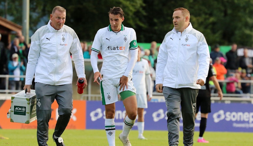 Florian Neuhaus (M.) verlässt gemeinsam mit Physiotherapeut Dirk Müller (l.) und Mannschaftsarzt Dr. Ralf Doyscher beim Testspiel von Borussia Mönchengladbach gegen den FC Ingolstadt am 26. Juli 2023 den Platz. Neuhaus' linker Oberschenkel ist bandagiert und er hält sich die Adduktoren.