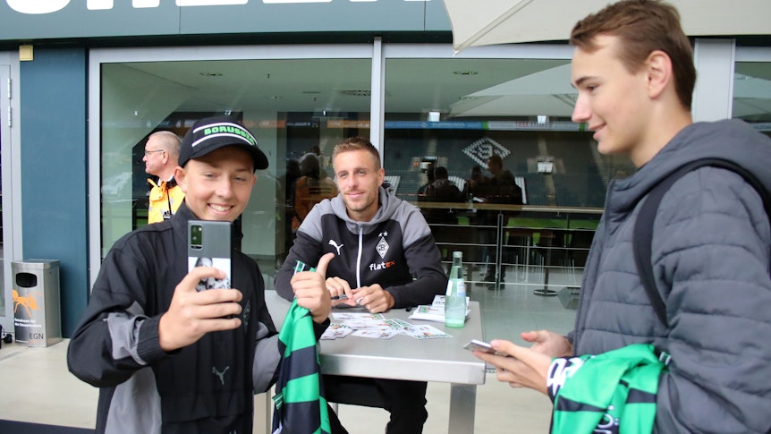 Herr Herrmann, ein Selfie bitte! Die Gladbach-Profis wie Patrick Herrmann haben sich zur Saisoneröffnung am Sonntag (6. August 2023) den Fans hautnah für Autogrammwünsche gestellt. Familienfest bei Borussia. Herrmann lächelt und sitzt an einem Tisch, derweil machen Fans gemeinsame Fotos.