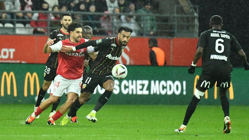 Reda Khadra im roten Reims-Trikot kämpft um den Ball mit Angelo Fulgini im schwarzen Lens-Trikot. Es regnet.