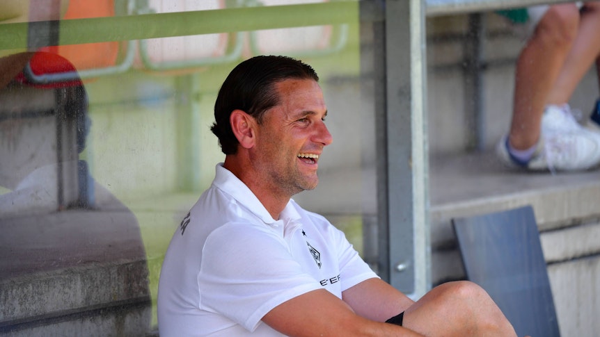 Gerardo Seoane sitzt auf der Trainerbank und lacht.