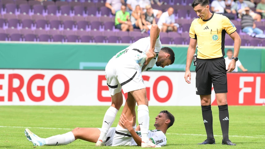 Borussia Mönchengladbachs Fabio Chiarodia liegt am Boden, Tim Kleindienst hilft ihm beim Dehnen. Schiedsrichter Florian Badstübner beobachtet die Szene.