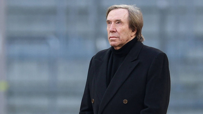 Günter Netzer steht in der Münchner Allianz-Arena auf dem Rasen und hat die Hände gefaltet.