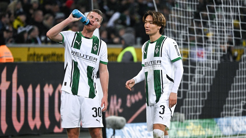 Die Gladbach-Profis Nico Elvedi und Ko Itakura stehen beim Spiel gegen Borussia Dortmund nebeneinander, Elvedi trinkt aus einer Wasserflasche.