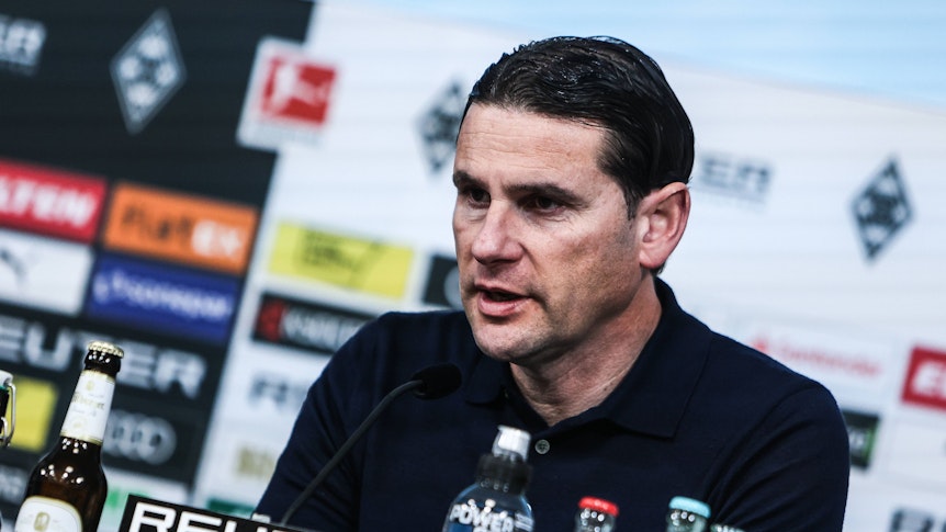 Borussia Mönchengladbachs Trainer sitzt bei der Pressekonferenz auf dem Podium und spricht ins Mikrofon.