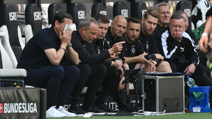 Blick auf die Bank von Borussia Mönchengladbach: Trainer Gerardo Seoane vergräbt sein Gesicht in einem Taschentuch, Roland Virkus schaut konzentriert auf das Spielfeld.
