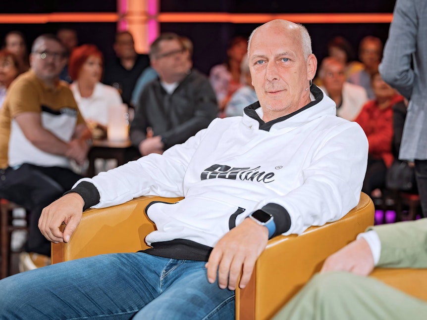 Mario Basler sitzt auf einem senffarbenen Sessel und hat beide Arme auf den Armlehnen liegen.