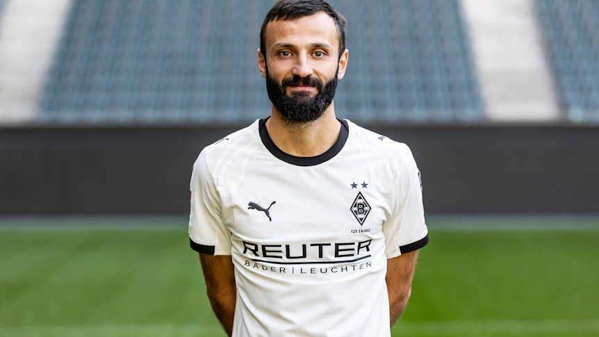 Porträt eines bärtigen Fußballspielers in weißem Gladbach-Trikot lächelnd vor Stadionhintergrund.