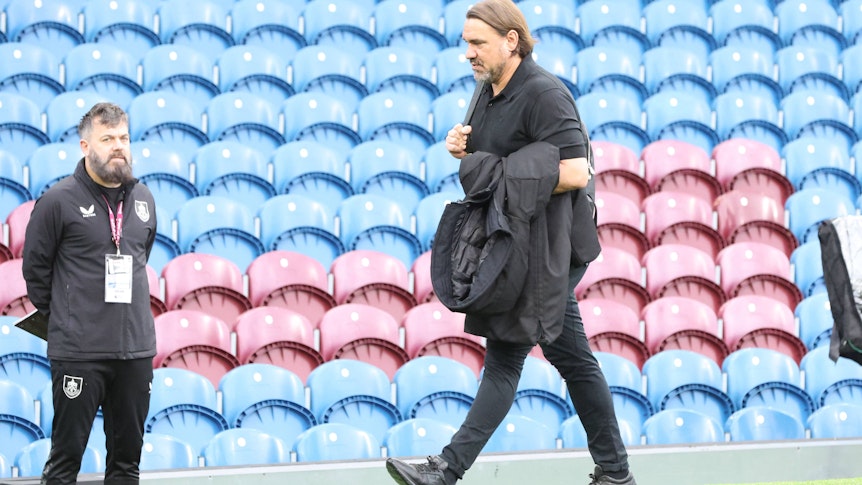 Daniel Farke geht mit einer Jacke vor blauen und roten Stadionsitzen entlang.