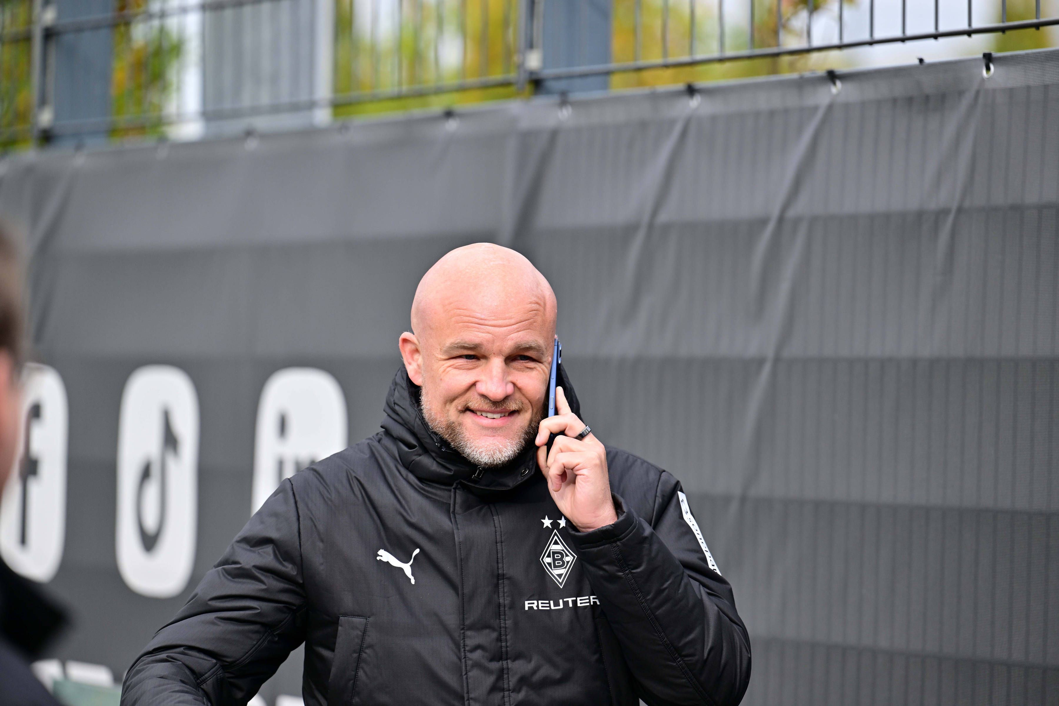 Rouven Schröder telefoniert in Gladbach-Jacke am Trainingsplatz und grinst dabei.